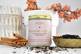 Cinnamon chai latte candle in a jar surrounded by cinnamon sticks, a bowl of black tea leaves, and an orange succulent plant. 