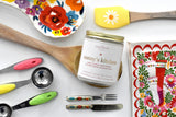 Nanny's kitchen candle in a jar on a wooden spoon surrounded by colourful and floral kitchen utensils.
